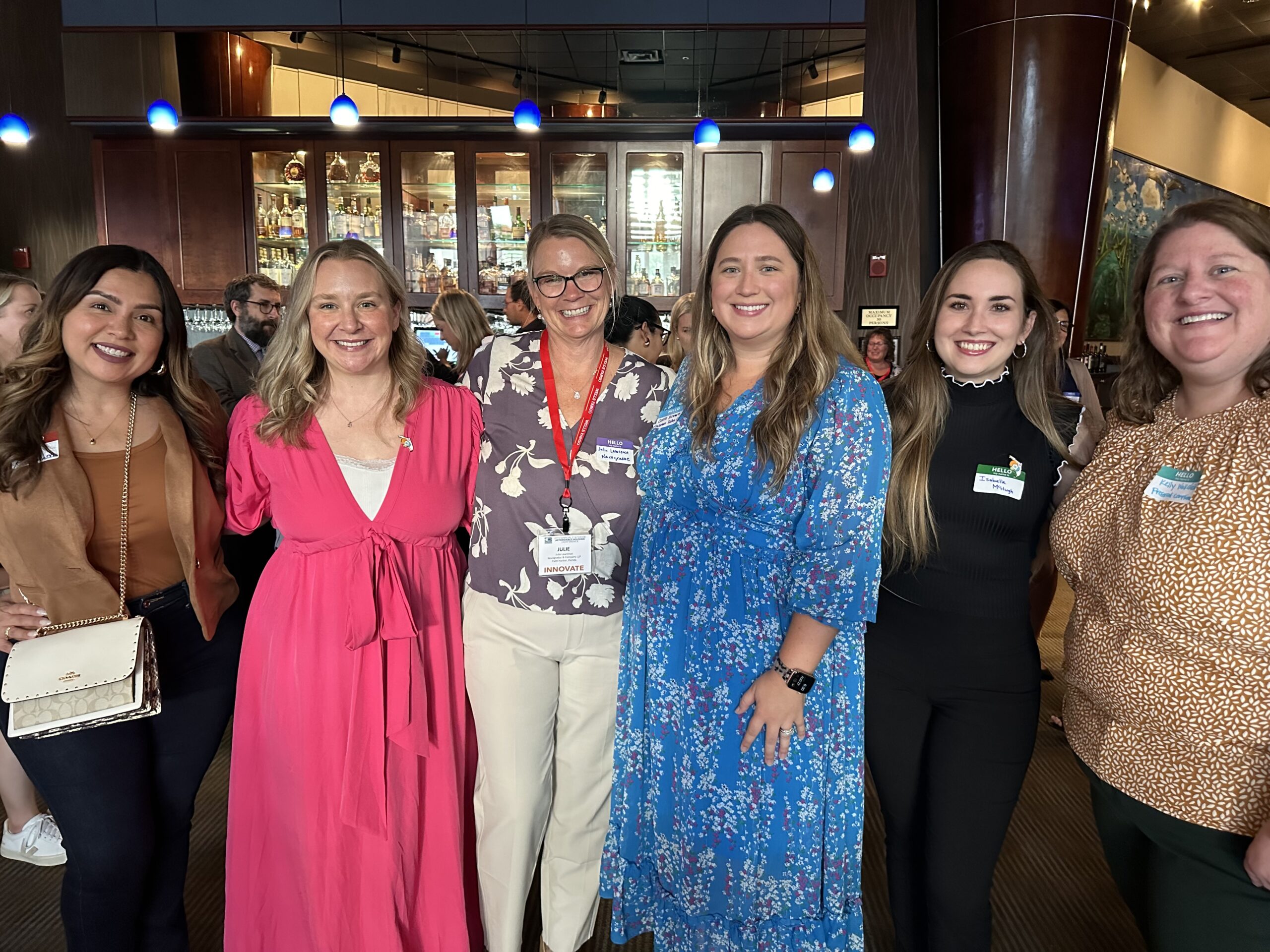 Women at the Florida Housing Coalitions Annual Affordable Housing Conference: Image of 6 women, from left to right, woman with brown hair, tan blazer, tan blouse and cream colored leather purse with gold chain strap, Hannah Anderson, sandy blonde hair, smiling, wearing bright pink dress with white camisole under. Woman with sandy blonde hair, hair pulled out of face with small pieces falling into her face, in floral pale purple blouse with white flowers on it wearing light khaki trousers, Cassie Brown, long brown hair, wearing long chiffon blue dress with small red and white flowers. Woman with dark brown hair wearing black blouse with white ruffle turtle neck collar wearing black trousers. Woman with smile, brown hair, tan shirt with spots of white and dark green trousers.